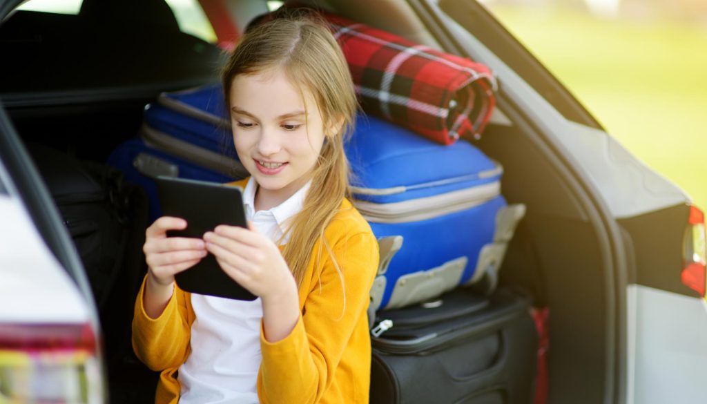 Girl reading a Kindle e-Reader sitting in open back of station wagon with camping gear packed in it.