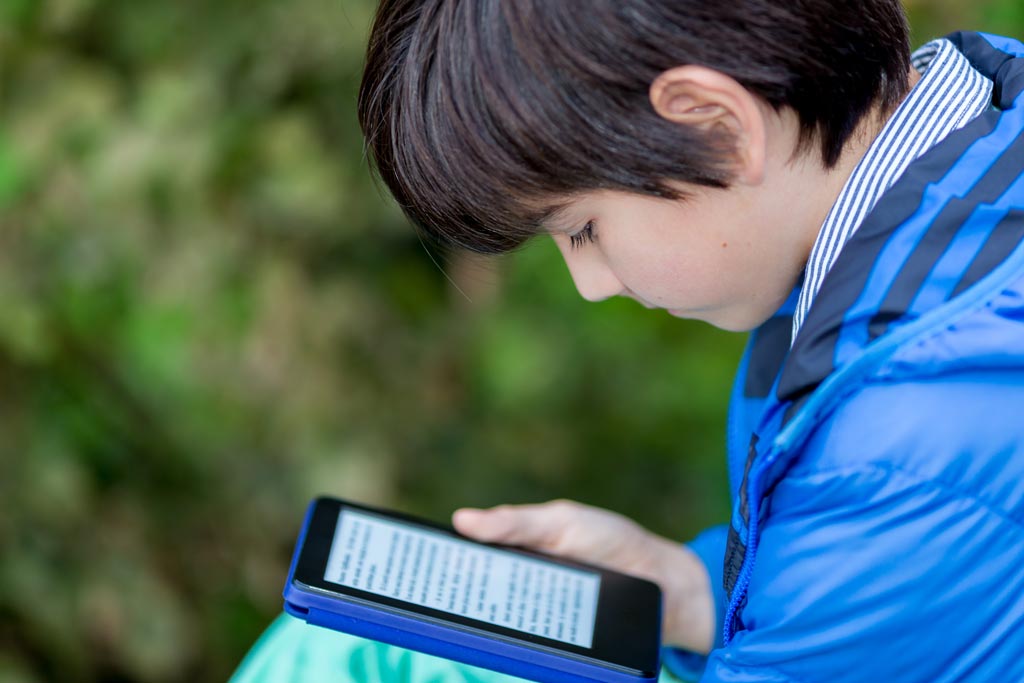 Middle school-age boy in blue jacket reading intently on Kindle e-reader oustide